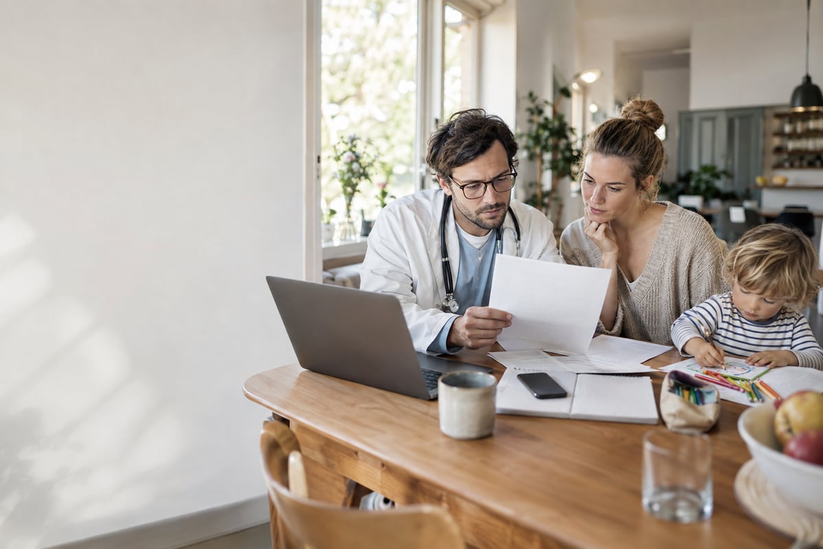 Arzt mit Partnerin und Kind an einem Tisch mit Laptop und Unterlagen